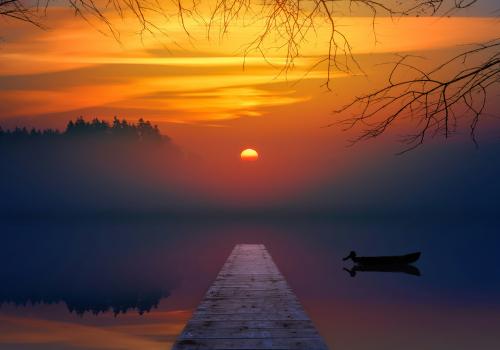 Sunset over a serene lake with a pier and silhouetted boat, hues of orange and blue.