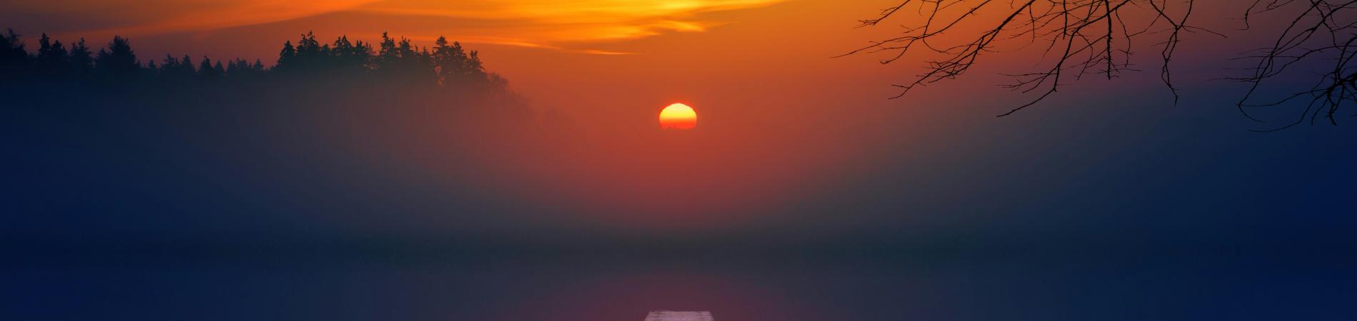 Sunset over a serene lake with a pier and silhouetted boat, hues of orange and blue.