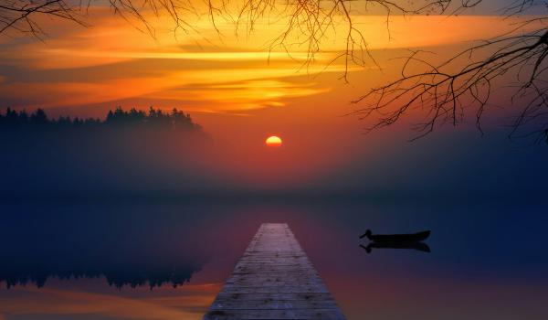 Sunset over a serene lake with a pier and silhouetted boat, hues of orange and blue.